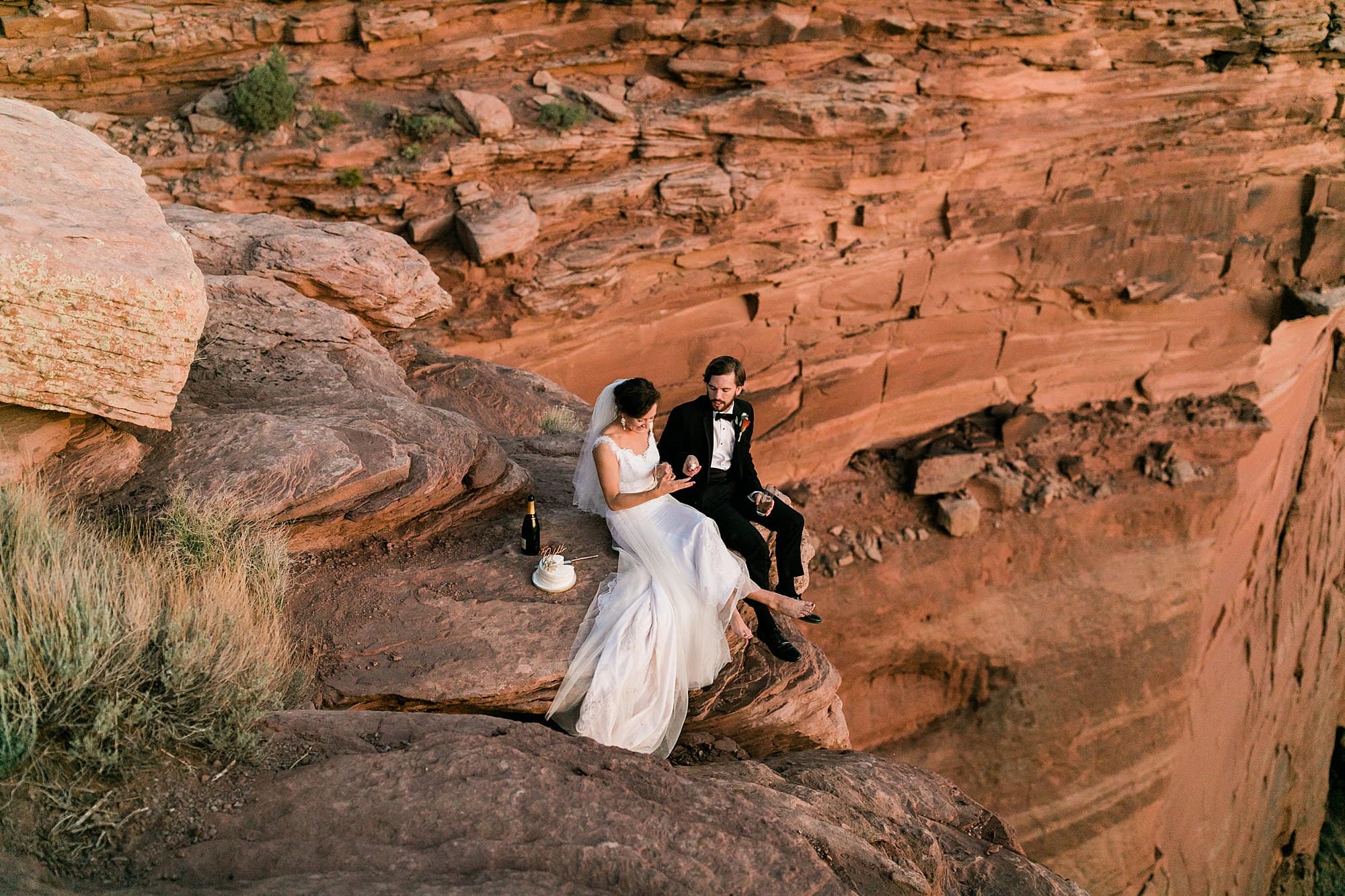 A couple sits on a cliff's edge and shares champagne and cake in Moab, Utah.