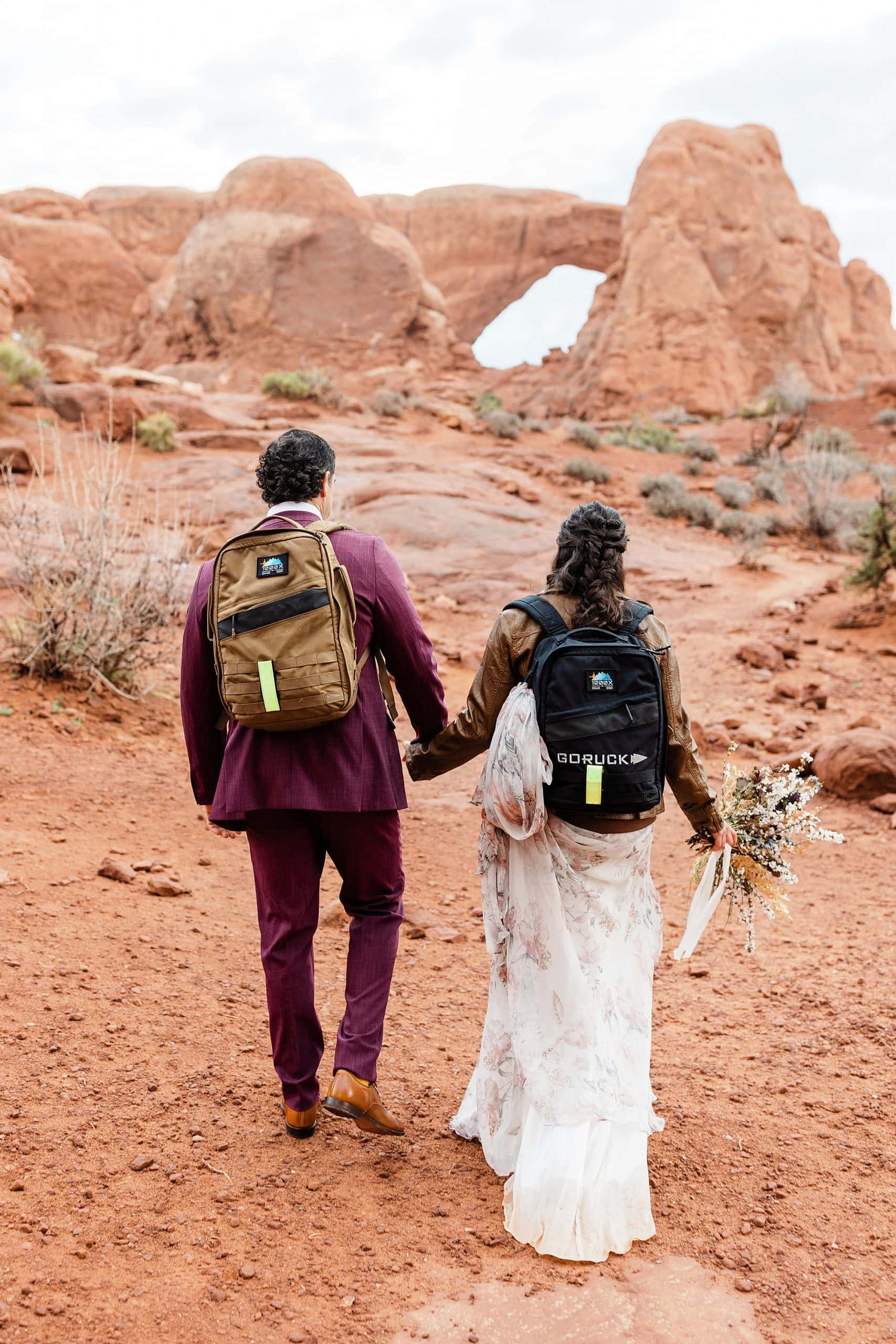 A couple wearing backpacks hikes to their ceremony location before they elope in Moab.