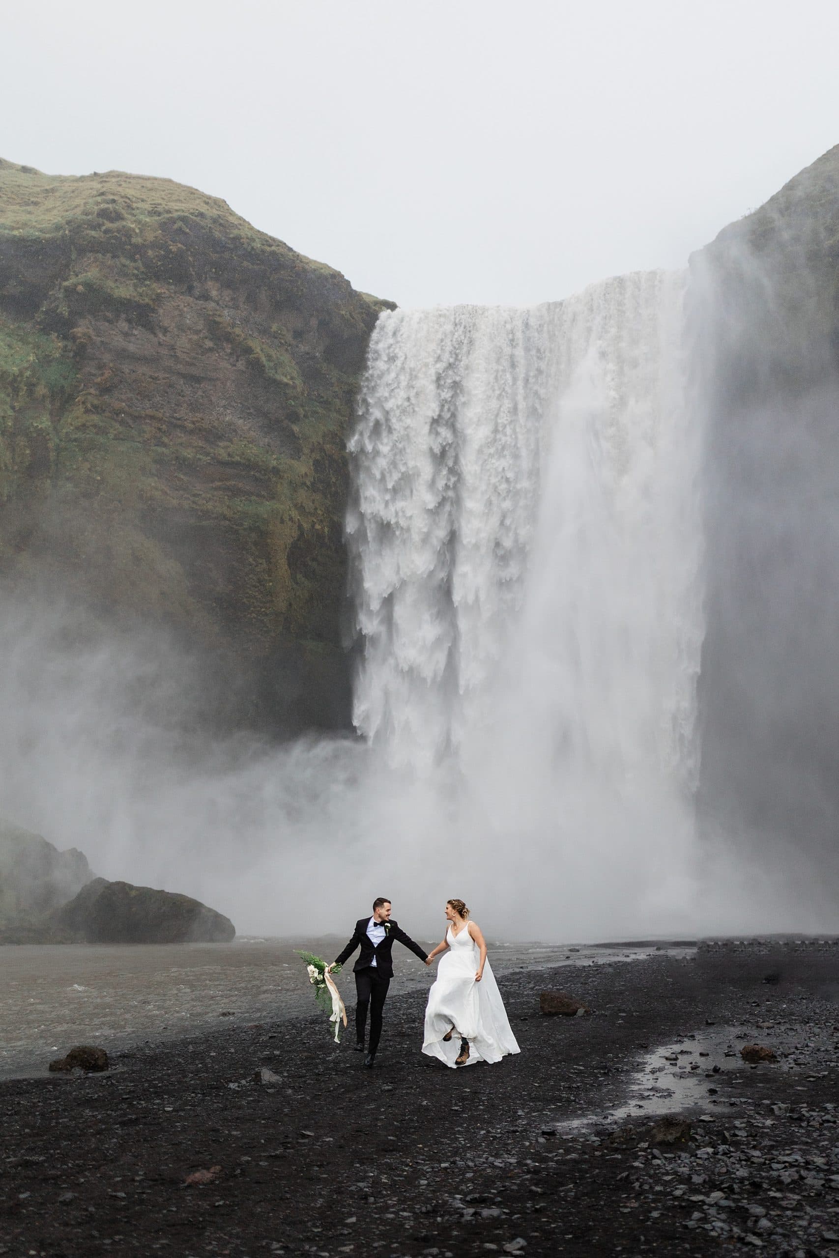 I couple skips while holding hands in front of Skógafoss after they elope in Iceland