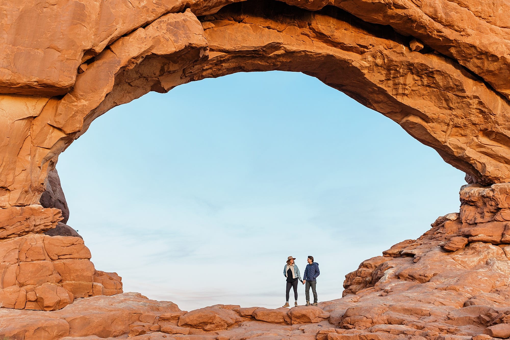 An elopement photographer and elopement videographer stand beneath a red rock arch in Moab, Utah. 