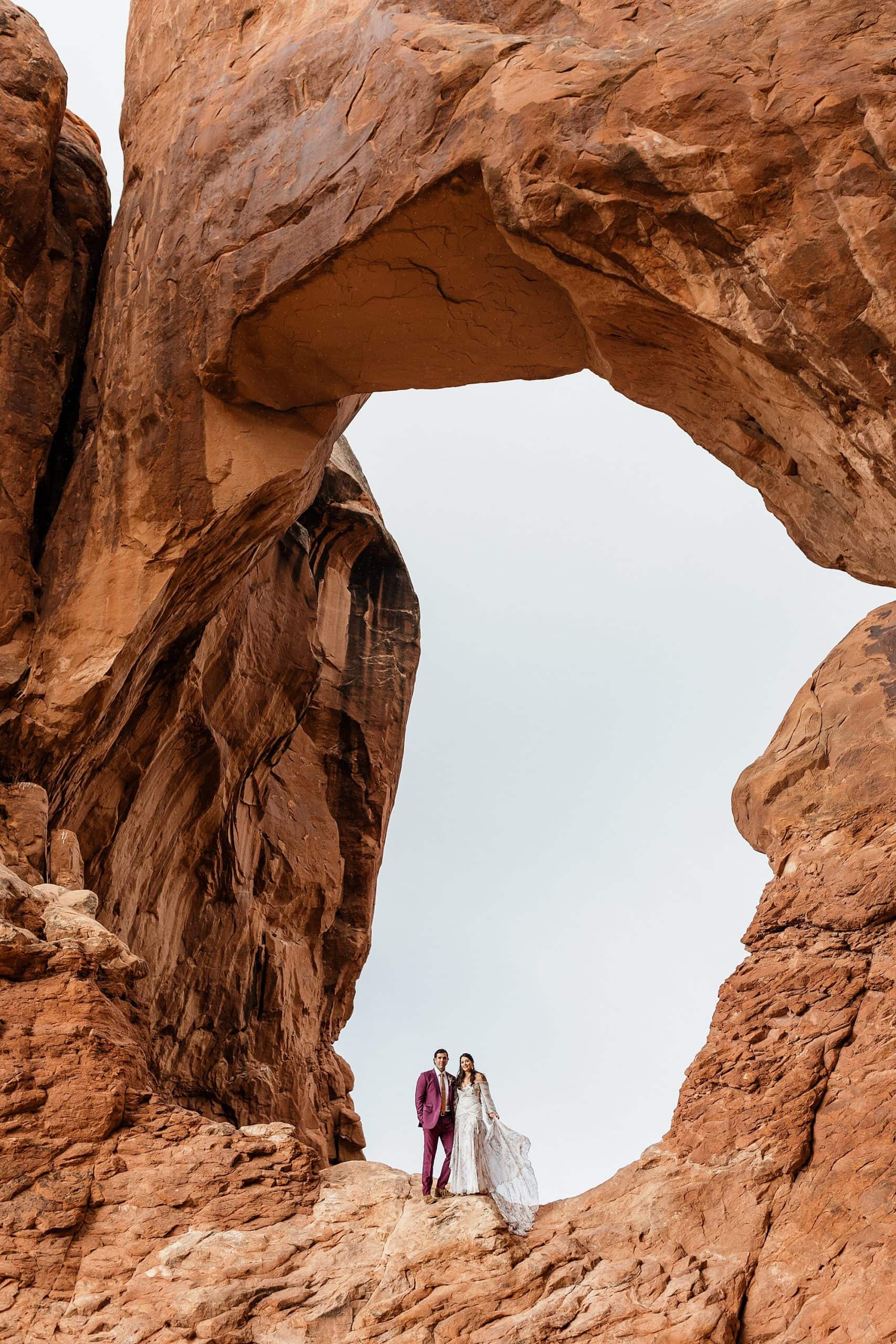 A couple stands beneath a red rock arch while they elope in moab.