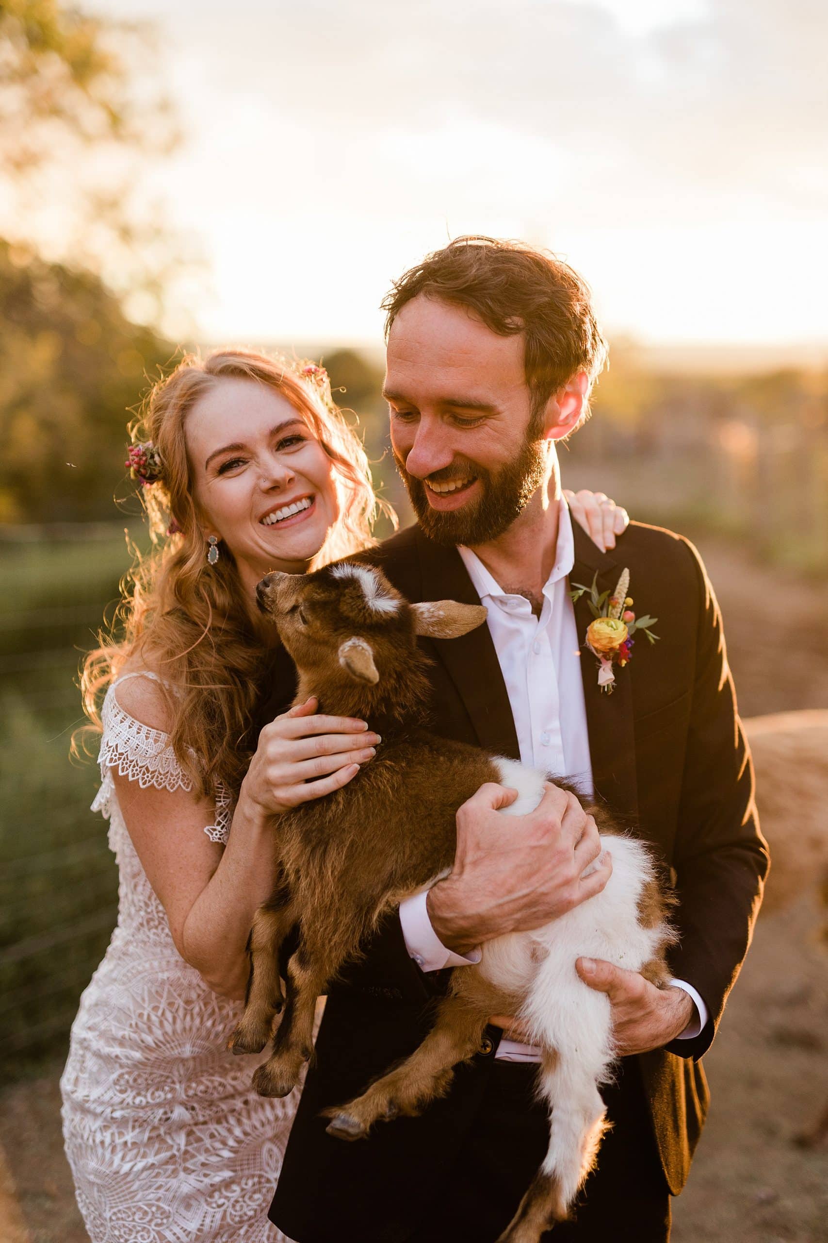 A couple smiles and laughs while holding a goat after their intimate wedding at a goji berry farm in Taos