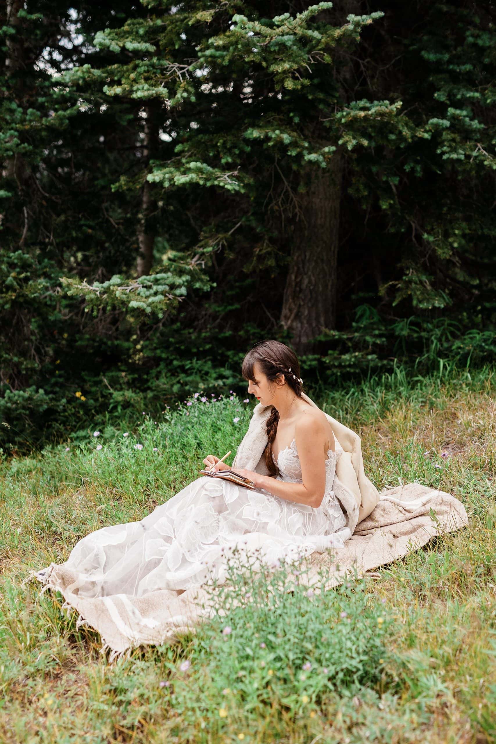 A woman in a wedding dress sits in a meadow before her elopement and writes in a vow book